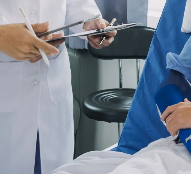 A patient in a hospital bed with a blue gown, with an arm in a cast, is talking to a doctor in a white coat who is taking notes on a clipboard.