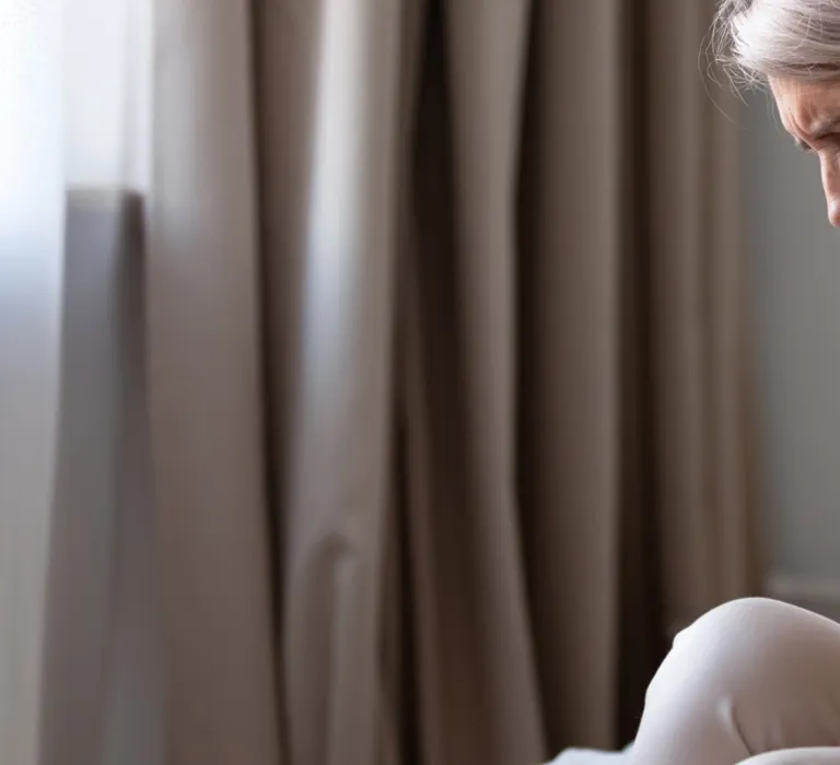 An older woman with gray hair sitting on a bed, holding her stomach and appearing to be in pain, in a softly lit bedroom with curtains and a small table in the background.