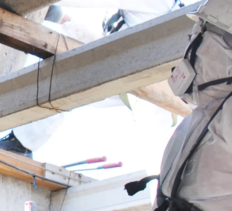 A construction worker in a white protective suit and helmet is working on a building's framework, with wooden and concrete elements, and safety equipment nearby.