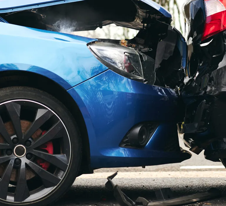 Two cars have collided head-on; the front of a blue car is crumpled against the rear of a black car, with visible damage and debris on the ground.