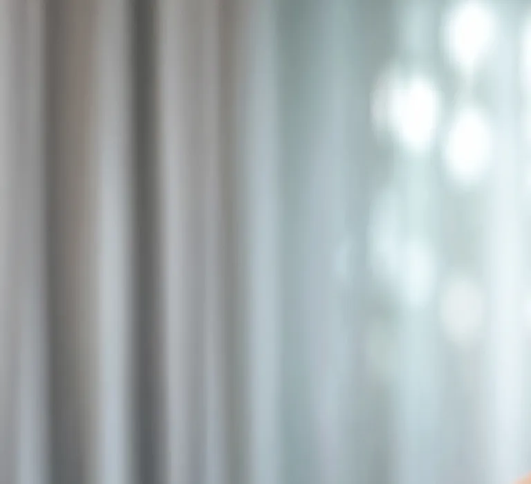 A man with a beard, holding his ear with a pained expression, standing in a softly lit room with curtains in the background.