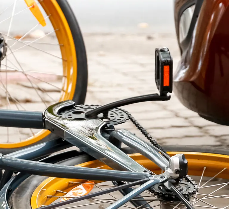 A fallen bicycle with yellow wheels lies on a cobblestone ground next to a parked car with black and gray alloy wheels.