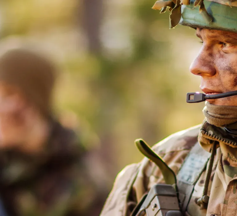 A soldier in camouflage gear, wearing a helmet and communication headset, standing outdoors with a blurred forest background.