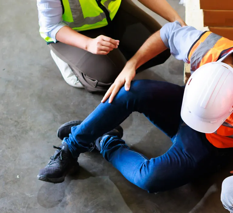 Three construction workers assisting an injured person lying on the ground, all wearing safety helmets and high-visibility vests, indoors near wooden pallets.