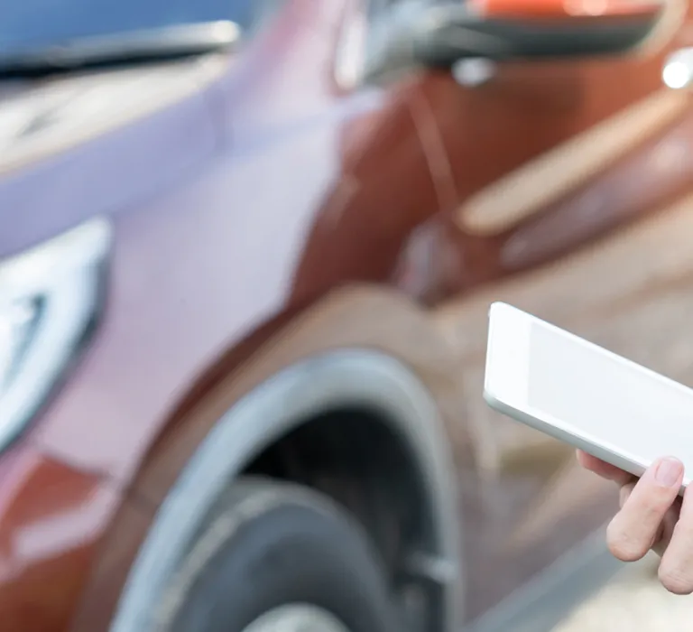 Two people are discussing a red car, with one holding a tablet and the other pointing at it, while standing outdoors near the vehicle.