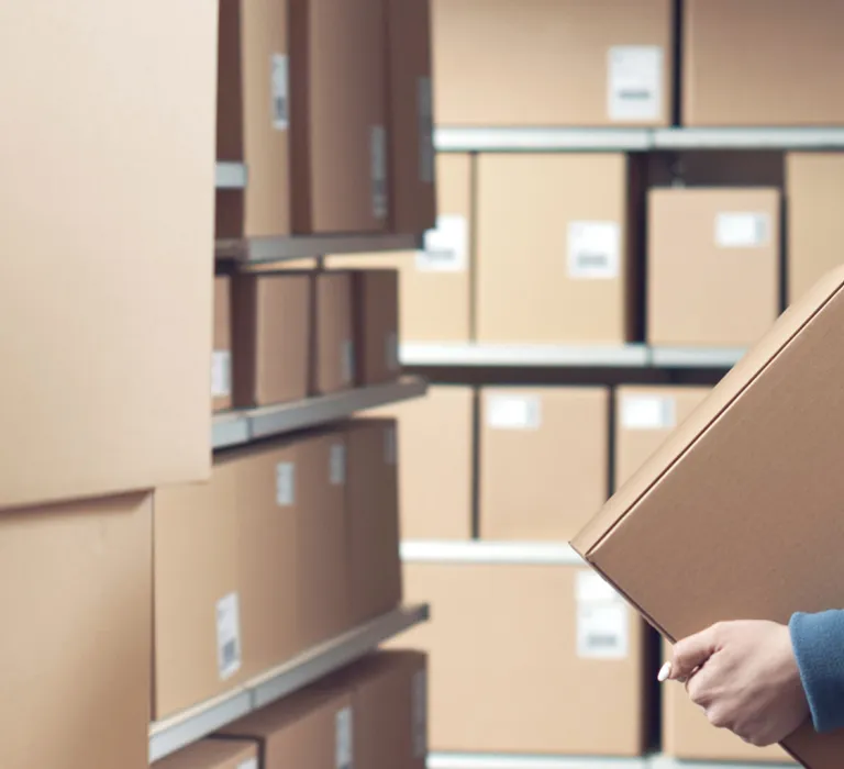 A woman in a blue shirt and orange safety vest is holding a cardboard box in a warehouse with shelves full of labeled boxes.