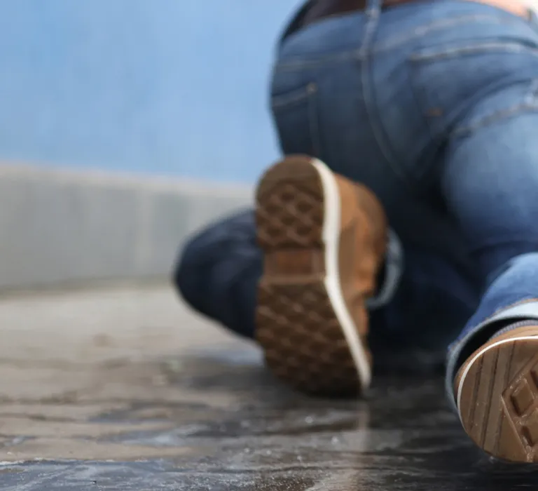 A person in jeans and brown shoes is crawling on a wet sidewalk near a backpack, with a blue wall and blurred pedestrians in the background.