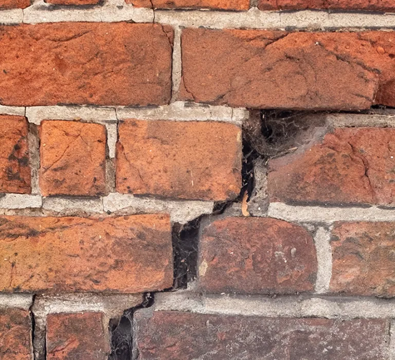 A close-up of a brick wall with a section of old, weathered bricks cracked and uneven, next to rough concrete with pebbles embedded in it, and a white painted wooden post on the left.