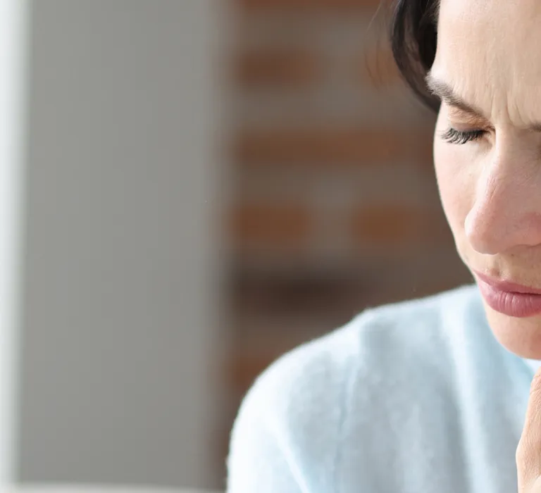 A woman with short dark hair is touching her jawline with her hand, appearing to experience pain or discomfort, in a well-lit room with blurred background elements.