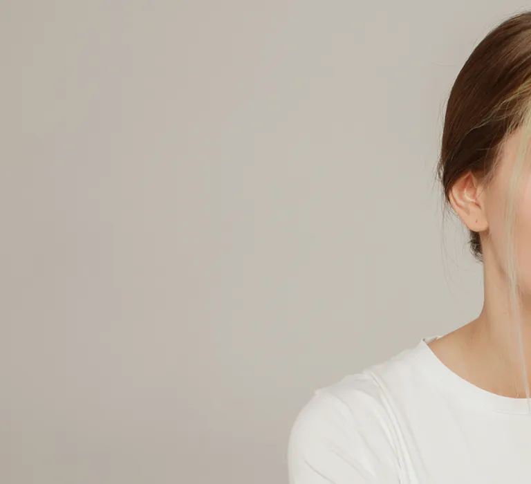 A woman with long hair and a white shirt looks at her reflection in a small mirror while wearing adhesive strips and a nasal patch on her nose.