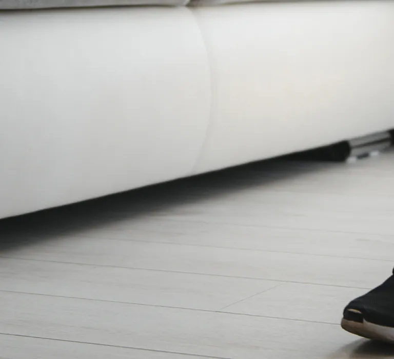 A person with a prosthetic leg is walking on a light-colored wooden floor near a white sofa, wearing black sneakers, black socks, and shorts.