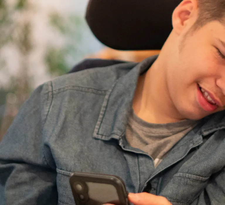 A young person in a wheelchair and an adult woman are smiling and looking at a smartphone together, sharing a moment of joy indoors with a blurred background.