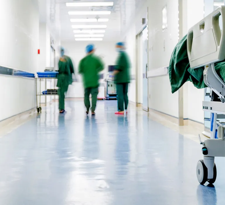 Empty hospital corridor with a wheelchair covered in green cloth on the right and medical staff walking away in the background.
