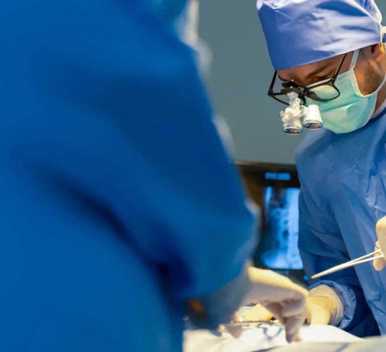 Surgeons in blue scrubs and masks perform surgery in an operating room, with bright lights overhead illuminating the procedure.