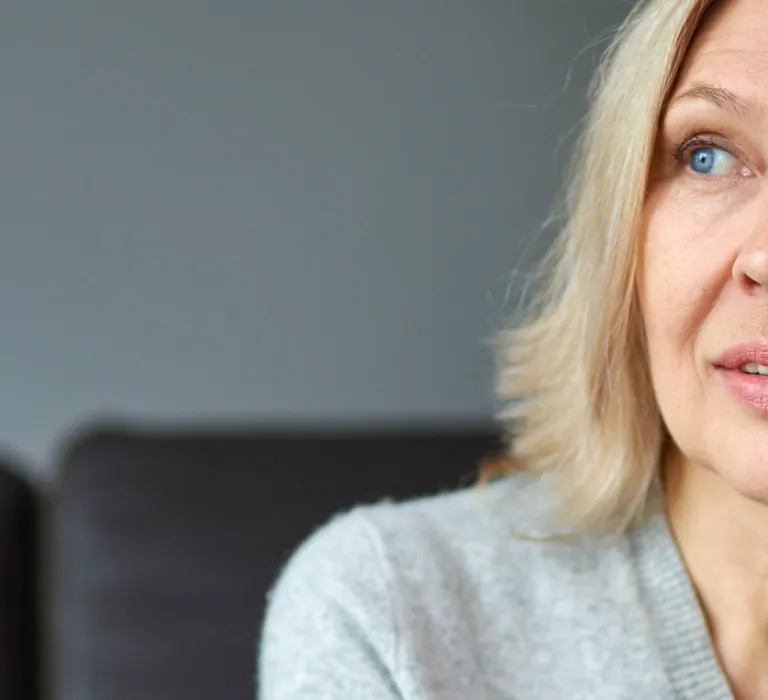 A mature woman with blonde hair and blue eyes is talking on a smartphone indoors, with a blurred green plant in the background.