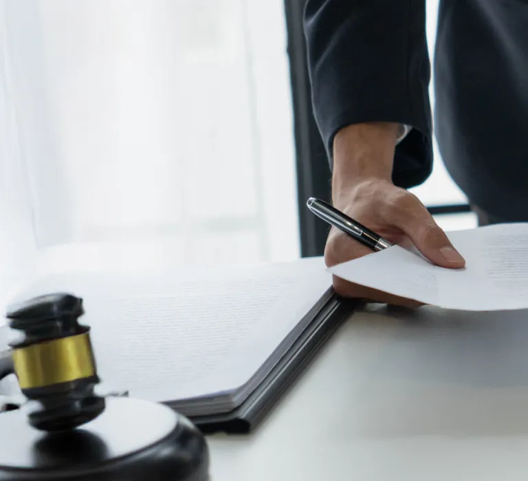 Person in a suit handling documents at a desk with a laptop, smartphone, and a wooden gavel.